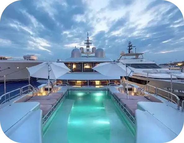 Big Boaty foredeck pool lit turquoise at dusk, with cushioned loungers on both sides, two white parasols and the superstructure behind
