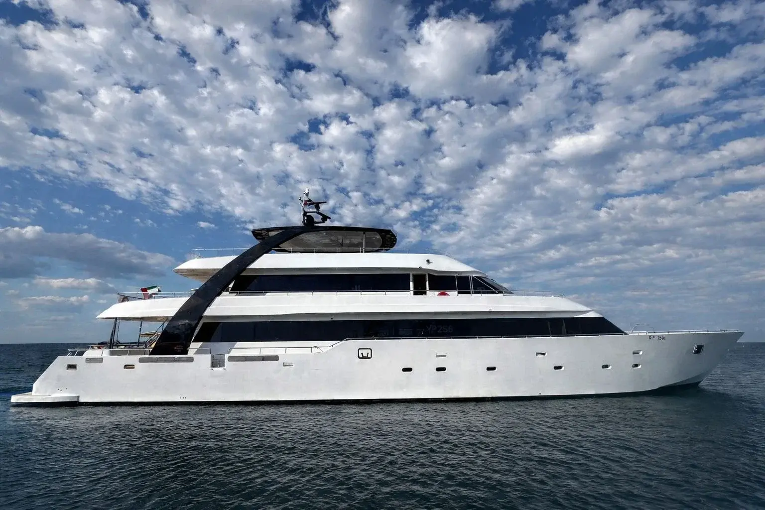 Full side profile of Carmen, a white 140 ft yacht with dark windows and a black arch over her upper decks, under a cloudy sky