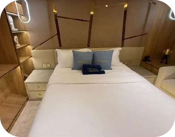 Cabin on I Don't Know with a white double bed, pale blue cushions, navy towels embroidered with the yacht's name and lit timber shelves