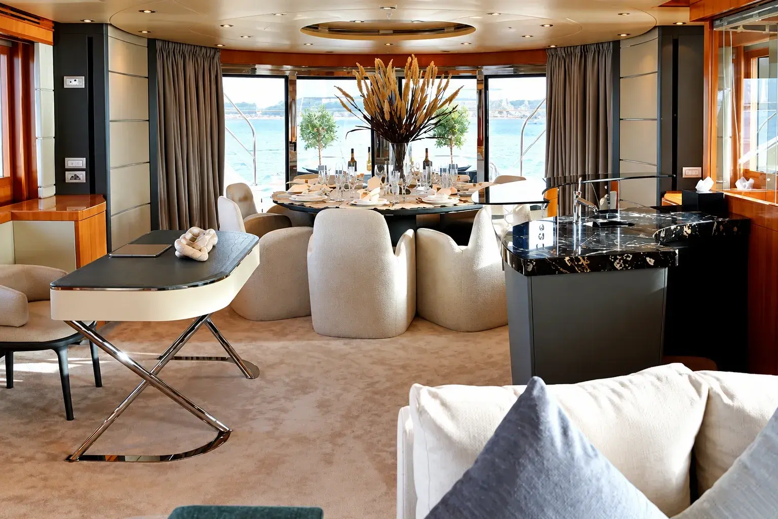 Illustration of the Royal Freedom saloon, with a round dining table set with glassware, cream chairs, a dark marble bar and glass doors open to the aft deck